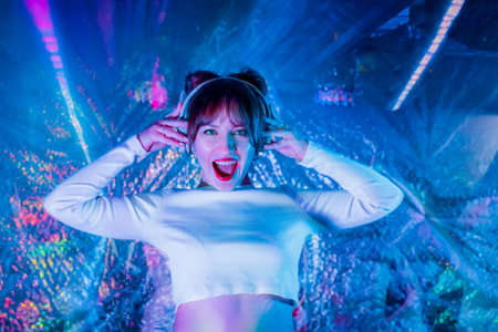Portrait of emotional screaming woman in white clothes and headphones dancing in neon light. Dj ,...
