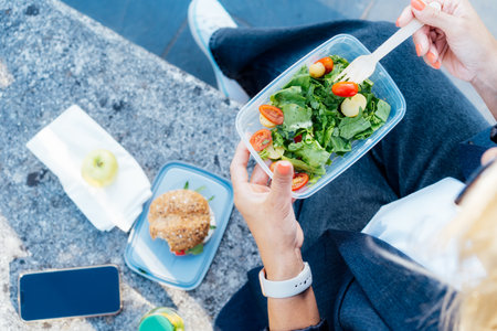 Top view close up business woman eating salad from her lunch box sitting on the bench at office park. Balanced diet lunchbox for weight loss. Healthy eating habits and well-being. Selective focusの写真素材