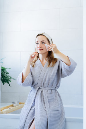 Young woman in bathrobe looking in mirror and making face massage with gua sha roller in bathroom. The concept of natural procedures for skin care. Home Beauty self-care treatment. Selective focus.の写真素材