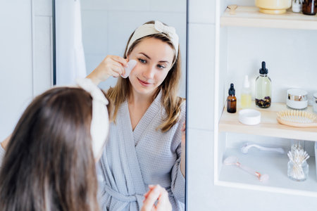 Young woman in bathrobe looking in mirror and making face massage with gua sha roller in bathroom. The concept of natural procedures for skin care. Home Beauty self-care treatment. Selective focus.の写真素材