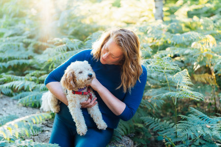 Happy middle-aged woman embracing her cockapoo puppy pet dog during walk outdoors in summer forest or park on sunset. Cute little doggy. A woman petting her dog like a small child. Selective focusの写真素材