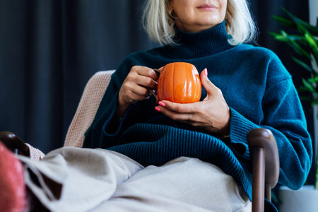 Cropped mature middle aged woman in sweater and warm socks relaxing in armchair with pumpkin shaped cup of hot coffee or tea drink. Cozy calm autumn holidays at home. Fall hygge mood conceptの写真素材