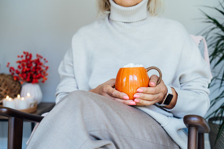 Close up pumpkin shaped cup of hot coffee drink with marshmallow in female hands with fall mood decor for hygge home on background. Cozy autumn beverage for Thanksgiving or Halloween holidaysの写真素材