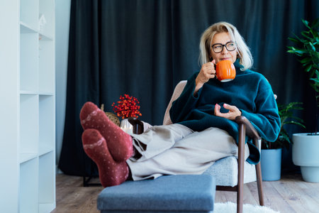 Middle aged woman relaxing with pumpkin shaped cup of hot drink in scandy style hygge interior home with fall mood decor. Lady dreaming, enjoy calm mood without stress, well being alone. Cozy autumn.の写真素材