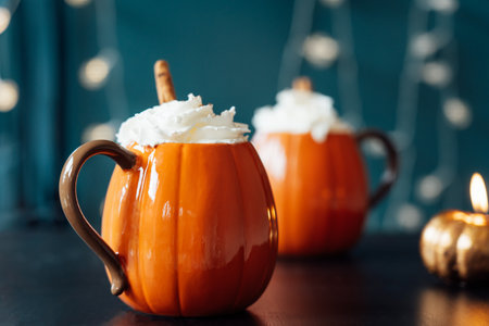 Pumpkin shaped cups of hot drink with whipped cream, cinnamon stick on black table with dark background. Pumpkin latte with spices. Cozy autumn beverage for Thanksgiving, Halloween holidays at homeの写真素材