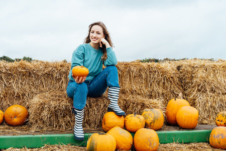 Smiling young woman sitting on straw bales and holding pumpkins. selecting best one for Thanksgiving and Halloween holidays decoration on agriculture farm. Pumpkin harvest. Autumn fall festive moodの写真素材