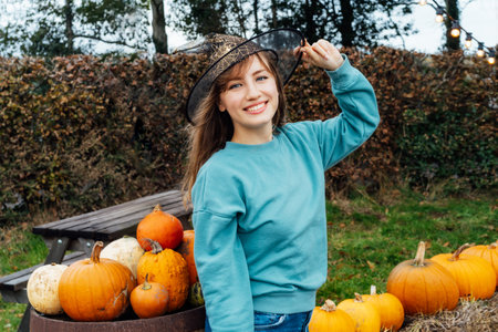 Portrait of smiling young woman in witch hat posing on pumpkins background. Female selecting best...