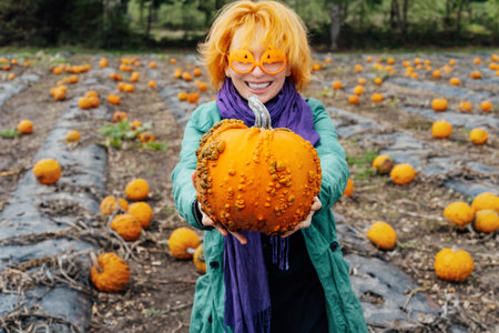 Focus on ugly pumpkin on the hands of emotional red head young woman in funny glasses on pumpkin patch field. Selecting best one for Thanksgiving and Halloween holidays. Autumn positive festive moodの写真素材