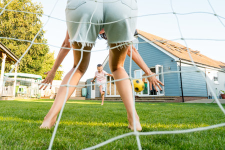 Kid kicking football ball while playing with mom, who stands on net on Backyard Lawn on Sunny Summer Day. Active family having fun outdoors enjoying leisure time. Childhood, happy lifestyle conceptの写真素材