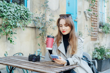 Female traveler chatting, sharing files, resting in cafe terrace table with vintage camera and coffee in reusable cup after city walk. Girl using internet in wifi zone, enjoying coffee and leisureの写真素材