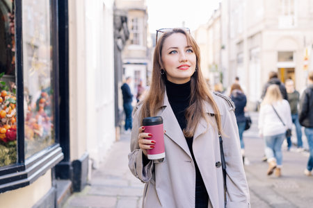 Young smiling woman in trench coat enjoying hot coffee drink in reusable cup while walking on the city streets decorated for the autumn holidays. Happy fall cozy mood, seasonal fashion style trend.の写真素材