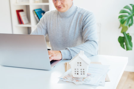 Focus on House model, money and docs on table and blurred senior man works on laptop. Housing cost and payments. Real estate, property investment. Home mortgage loan rate. Saving money for retirementの写真素材