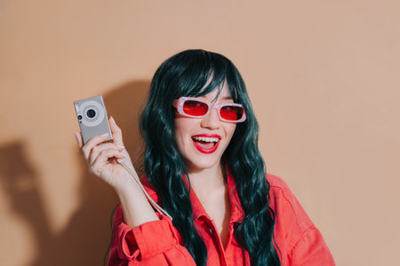 Cheeky shouting bright fashion girl with dark green long hair, bright red lips, top and sunglasses with compact digital camera on beige background. Trendy young hipster photographer, travel photoの写真素材