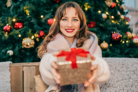 Merry Christmas and Happy Holidays. Beautiful young woman looking at camera and holding out Christmas gift box on background of a decorated Christmas tree. Exchanging presents. Festive mood.の写真素材
