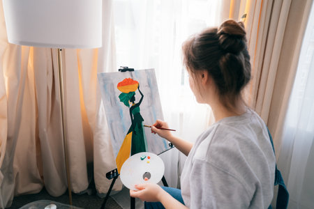 Back view female artist sitting in front of easel, painting picture with inspiration, drawing on canvas with paintbrush, palette at home. Art, hobby, leisure and creative occupation concept.の写真素材