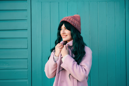 Stylish young smiling hipster woman with color hair wearing pink coat and knitted hat on turquoise green wooden background. Urban seasonal city street fashion. Barbiecore style. Selective focus.の写真素材