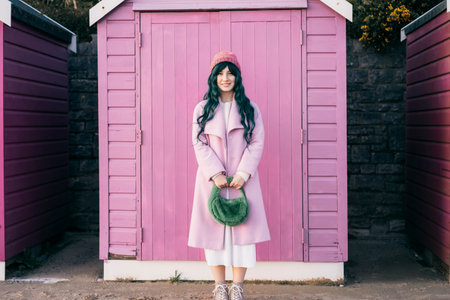 Stylish young smiling hipster woman with color hair wearing pink coat, knitted hat and fur bag on pink wooden beach hut background. Seasonal city street fashion. Barbiecore style. Selective focus.の写真素材