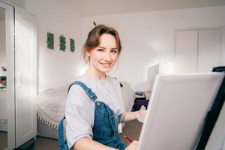 female artist sitting in front of easel, painting picture with inspiration, drawing on canvas with paintbrush, palette, smiling, relaxing at home. Art, hobby, leisure and creative occupation conceptの写真素材