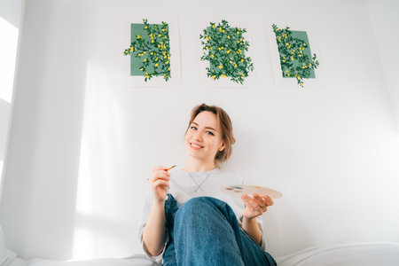 Smiling female artist sitting on bed with paintbrush, palette under her hand-drawn painting art works on the wall at home. Art, hobby, leisure and creative occupation concept. DIY, Home decorating.の写真素材