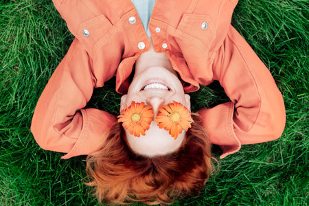 Top view happy smiling redhead Woman in trendy peach color jacket and gerbera Flower glasses lying on green grass. Positive Emotion people. Color of the 2024 year. Spring, summer moodの写真素材