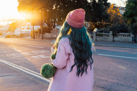 Back view stylish hipster woman with color hair walking the street at sunset in warm outfit wearing pink coat, knitted hat and fur bag, happy mood, seasonal fashion style trend. Selective focus.の写真素材