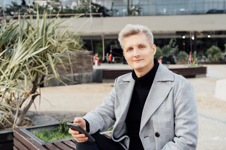 Portrait of young Handsome stylish man in elegant trendy coat using mobile phone while sitting on the bench in urban city background. Seasonal street fashion. Online communication, remote work.の写真素材