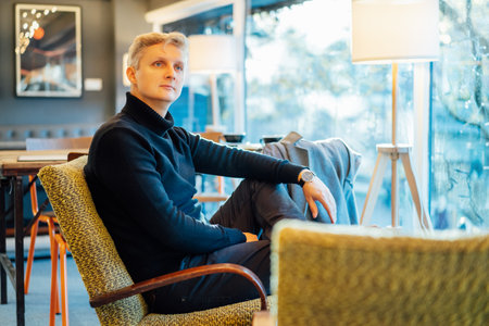 Portrait of thoughtful handsome young stylish man in black outfit relaxing on the vintage style armchair in modern cafe. Elegant fashion style. Vintage interior details. Selective focus.の写真素材