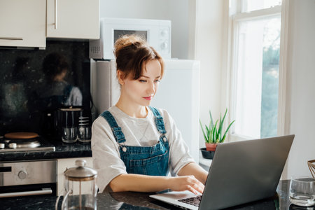 Remote work at home. Concentrated young woman working on Laptop at the Kitchen Table in tiny flat apartments. A female student having online classes, Freelancer working from home. Online Jobの写真素材