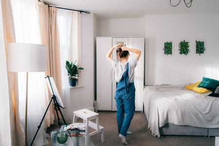 Back view female looking at the mirror and putting hair in a bun while planning to take up drawing class. Drawing on canvas. Relaxing at home. Art, hobby, leisure and creative occupation concept.の写真素材
