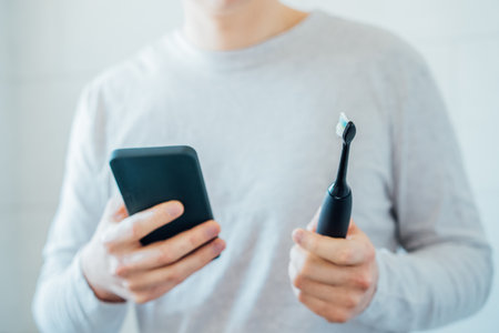 Close up man puts settings of his electronic toothbrush with mobile phone app. Wireless connecting sonic toothbrush with smart phone app. Modern home health care technology concept. Selective focusの写真素材
