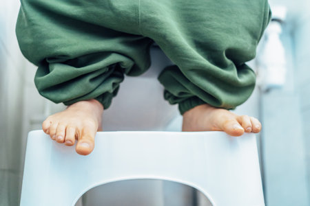 Close up adult male sitting on white toilet with his pants down and his feet are standing on toilet foot Stool for an easy process of bowel movements, going to the toilet to poop. Selective focus.の写真素材