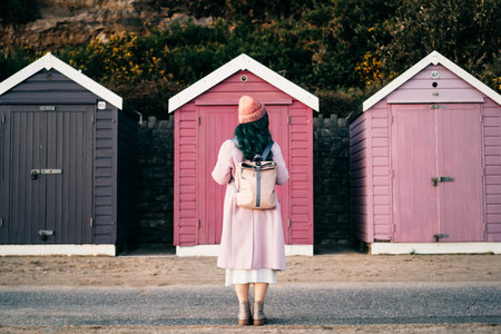Back view stylish hipster woman with color hair in total pink outfit and backpack looking at wooden beach huts. Off season Travel concept. Seasonal street fashion. Barbiecore style. Simple pleasuresの写真素材