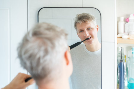 Young Man is using electric ultrasound toothbrush. Male brushing teeth and looking in mirror. Daily dental hygiene and oral health. Morning routine, young man cleaning his teeth. Selective focusの写真素材