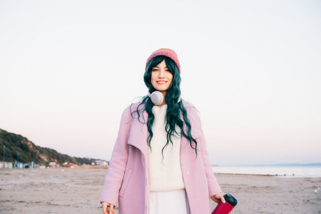 Stylish young smiling hipster woman with color hair wearing wireless headphones, pink coat, hat, walking on beach with reusable coffee cup. Off season Travel and freedom concept. Simple pleasuresの写真素材