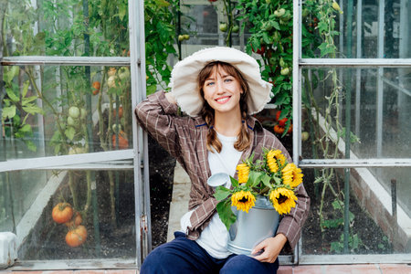 Portrait of smiling young female farmer woman holding watering can with fresh sunflowers bouquet, sits near greenhouse with ripe tomatoes. Urban farming lifestyle. Growing organic vegetables in gardenの写真素材