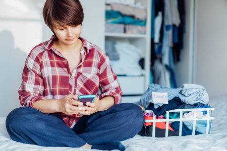 Closeup woman using smartphone to find places where to donate or sale clothes. Decluttering, Sorting clothes and Cleaning Up. Conscious consumer, sustainability. Reuse, second hand, Charity conceptの写真素材
