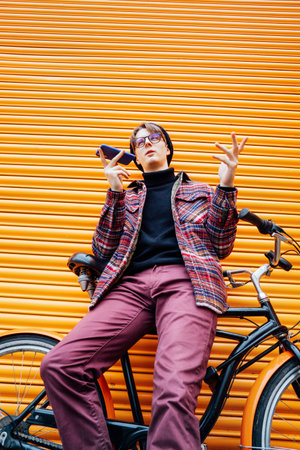 Young hipster man recording voice message on phone while sitting on cruiser bicycle near orange...