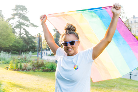 Mixed race woman raising a rainbow flag outdoor. Young lesbian activist smiling and holding flag symbol of social movement Lgbtq. Fight for equality, freedom and human rights. Celebrating Pride month.の写真素材