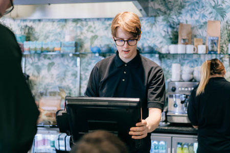 Young waiter serving customer at cash point in cafe. Man working with POS terminal. Cashier, barista checking for payment receipt. Hospitality, server and preparing a slip at the till in coffee shop.の写真素材