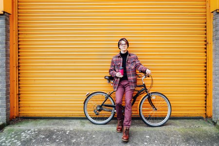 Young smiling hipster male student enjoying coffee out of reusable thermos cup while sitting on cruiser bicycle near orange wall. Eco friendly transport for carbon footprint. Sustainable lifestyle.の写真素材