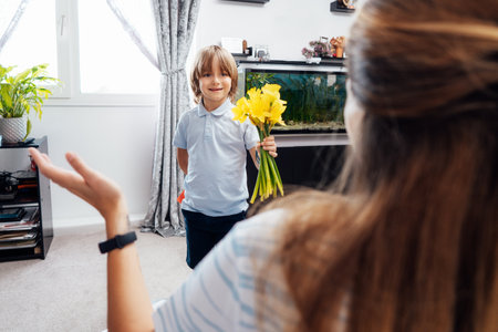 Smiling boy holding flower bouquet and self made greeting card for mother behind back while making surprise. Happy mothers day. Child boy congratulations mom and gives her flowers and postcard.の写真素材