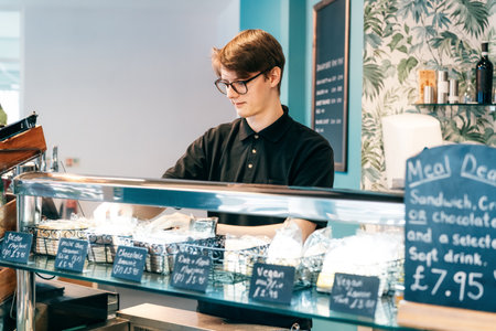 A young waiter displays the goods in a modern coffee shop counter. Student working part-time in cafe. Problems of unemployment and Job opportunities for youth. Small business vacanciesの写真素材