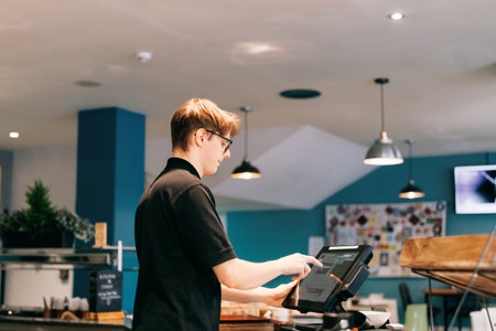 Young waiter serving customer at cash point in cafe. Man working with POS terminal. Cashier, barista checking for payment receipt. Hospitality, server and preparing a slip at the till in coffee shopの写真素材