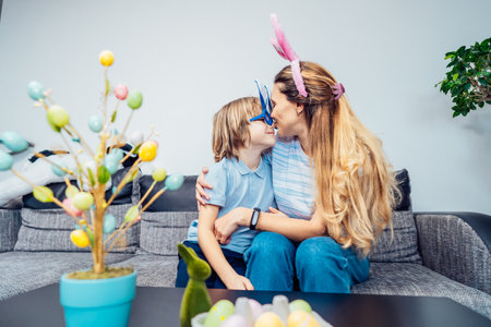 Happy smiling Caucasian mother and her son in bunny ears having fun at home on Easter day. Family sitting on sofa at home hugging, mother kissing son. Family Easter traditions. Selective focusの写真素材