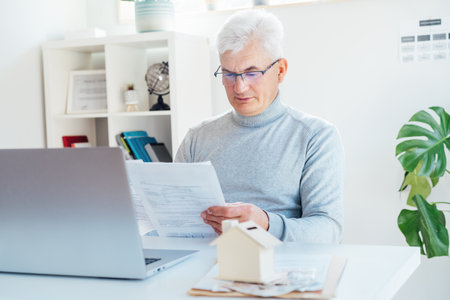 Middle aged man working on laptop with house model money box and docs on table. Housing cost and payments. Real estate, property investment. Home mortgage loan rate. Saving money for retirement.の写真素材
