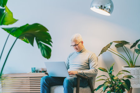 Concentrated gray hair middle-aged man working on laptop while sitting in armchair at home. Confident, experienced senior male professional. Small business entrepreneur manage business at home.の写真素材