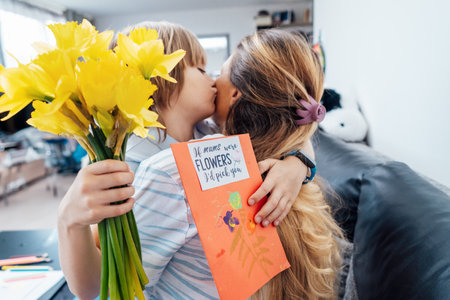Happy mothers day. Child boy hugging and kissing his mother after he congratulates mom and gives her flowers bouquet and handmade postcard. Family holiday and togetherness. Selective focus on card.の写真素材