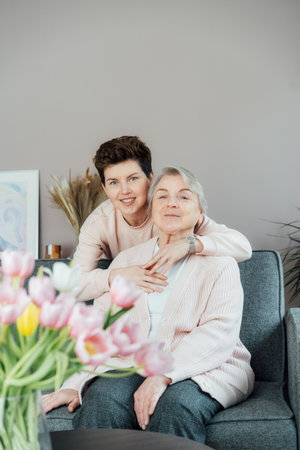 Senior 70s mother and adult neutral gender daughter hugging. Happy family enjoying weekend together in living room. Elderly woman spending quality time with daughter who visits her in retiring house.の写真素材