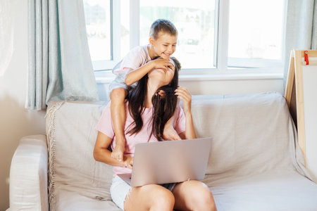 Young mother trying to work on laptop at home while her son wants to play. Childcare and working mom concept. Child make noise and disturb a woman at work. Homeschooling and freelance work.の写真素材