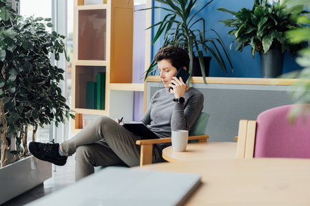 Confident smiling middle aged woman in casual clothing having conversation by phone, making note in notebook in office, meeting room. Phone interview. HR specialist, hiring, recruiting.の写真素材